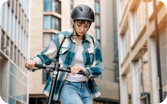 Sieviete ar melnu ķiveri, rūtainu krekliņu un džinsiem brauc ar elektrisko skrejriteni pilsētā, fonā augstu ēku logi.