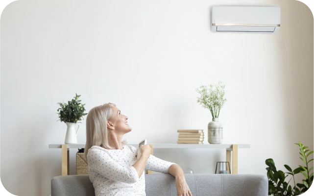 Sieviete sēž uz pelēka dīvāna dzīvojamā istabā un ar pulti rokā ieslēdz sienas gaisa kondicionieri; fonā plaukts ar grāmatām un divām vāzēm.