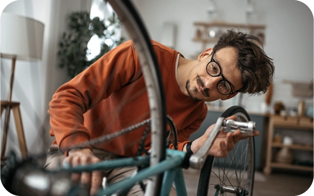 Vīrietis oranžā džemperī remontē zilas krāsas velosipēdu, kas ir nolikts ar riepām uz augšu, dzīvojamā istabā, koncentrējoties uz aizmugurējo ratu un ķēdes zonu.
