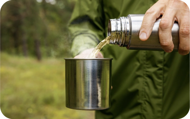 Cilvēks zaļā jakā lej kūpošu dzērienu no nerūsējošā tērauda termosa metāla krūzē; fonā koki un pļava.