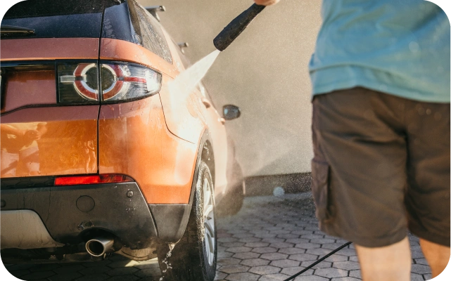 Vīrietis zilā kreklā un tumšos šortos ar augstspiediena mazgātāju tīra oranžu automašīnu, ūdens strūkla trāpa virsbūvei, mašīna novietota uz bruģētas virsmas.