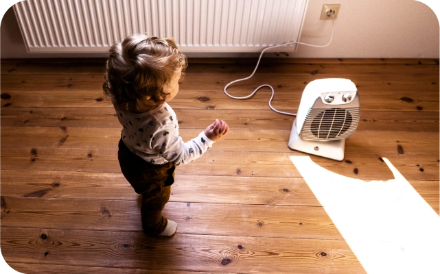 Mazs bērns stāv uz koka grīdas, pavērsis skatienu uz grīdas novietota maza, balta elektriskā sildītāja pusi, kas ir pieslēgta pie sienas rozetes; fonā pie sienas piestiprināts radiators.