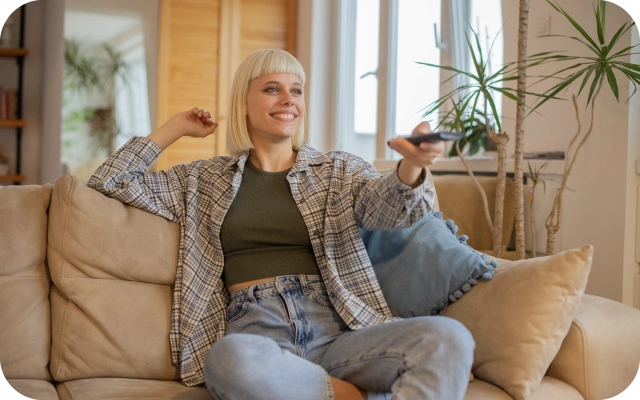 Sieviete sēž uz bēša dīvāna ar pulti rokā; fonā liels skapis ar spoguli un telpaugs ar šaurām lapām.