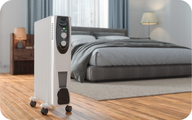 Balts eļļas radiators ar ritenīšiem stāv uz brūnas koka grīdas, fonā divguļamā gulta pelēkā krāsā, brūns naktsskapītis ar lampu, logs un pelēki nakts aizkari.