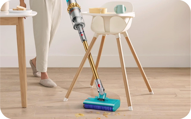 Persona gaišās biksēs izmanto zeltainu bezvadu putekļsūcēju, lai notīrītu koka grīdu no ēdiena drupačām ap bērnu krēsliņa koka kājām; fonā galds ar ēdienu.