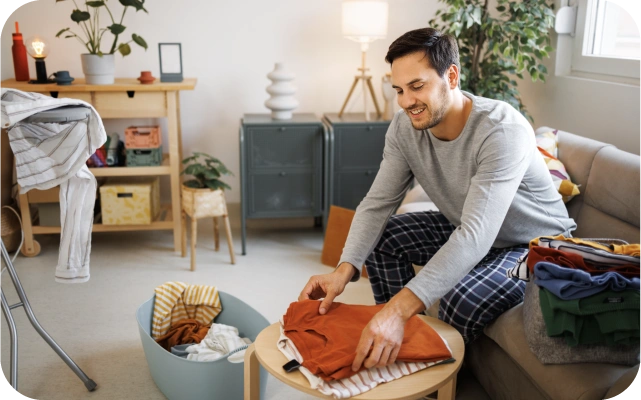 Vīrietis sēž viesistabā uz bēšas dīvāna un loka oranžu T-kreklu uz apaļa, koka kafijas galdiņa; blakus galdiņam ir zils veļas grozā ar veļu, fonā koka plaukts ar lampu un augu.