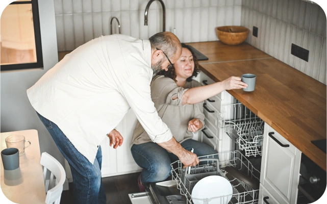 Vīrietis un sieviete virtuvē iztukšo trauku mazgājamo mašīnu; sieviete ir pietupusies un noliek krūzi uz galda virs trauku mazgājamās mašīnas, vīrietis ir noliecies virs atvērtās ierīces un izņemt virtuves piederumus.