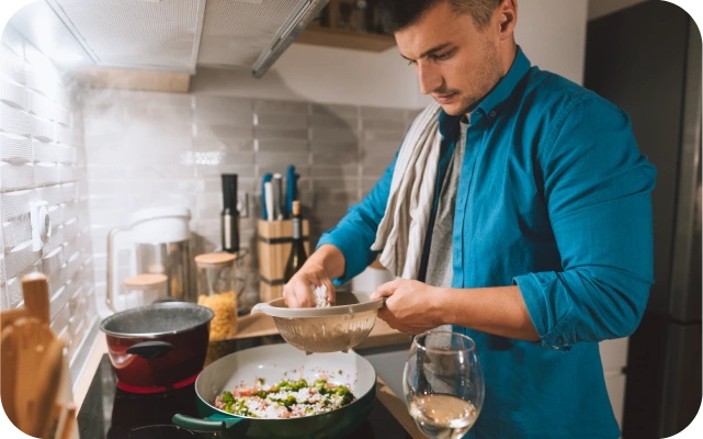 Vīrietis zilā kreklā virtuvē gatavo ēdienu — stāv pie pannas ar caurduri vienā rokā un ar otru roku izņem sastāvdaļas; fonā virtuves piederumi un glāze ar dzērienu.