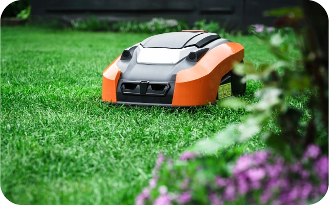 Oranžs un melns robots zāles pļāvējs darbojas zālienā; fonā ziedi un puķu dobes.