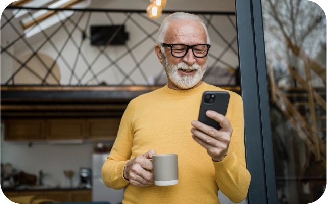 Vecāka gadagājuma vīrietis dzeltenā džemperī stāv atspiedies pret durvju aili, turot vienā rokā bēša krūzi, otrā — mobilo telefonu.