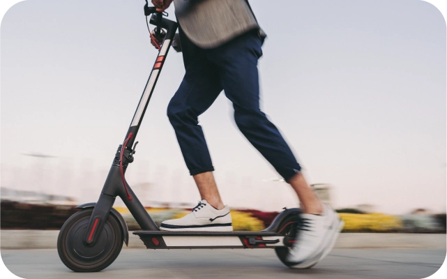 Cilvēks pelēkā jakā un tumšas biksēs brauc ar melnu elektrisko skrejriteni pilsētvidē, fonā neskaidras ēkas, radot kustības dinamiku.