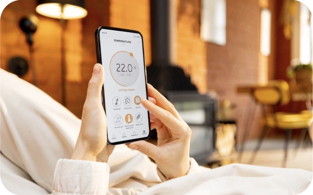 Cilvēks guļ uz dīvāna, turot viedtālruni ar atvērtu temperatūras kontroles lietotni, kurā iestatīta 22 °C temperatūra viesistabā.