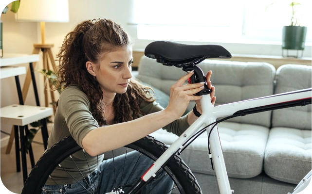 Sieviete pielāgo balta velosipēda sēdekli, izmantojot abas rokas, lai pievilktu sēdekļa stiprinājumu; fonā pelēks dīvāns, lampa un rakstāmgalds.