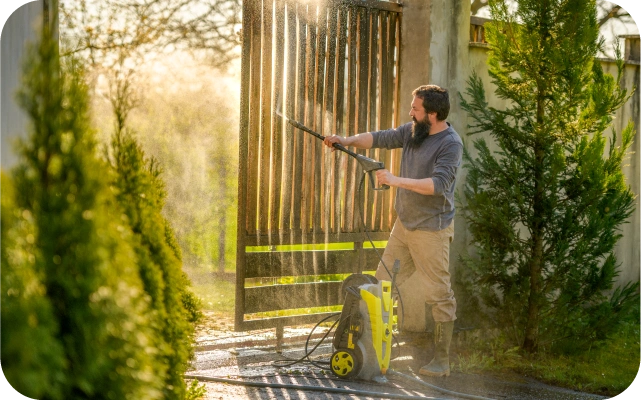 Vīrietis ar augstspiediena mazgātāju tīra koka vārtus, ūdens strūkla atspīd saulē, fonā zaļi krūmi un koki.