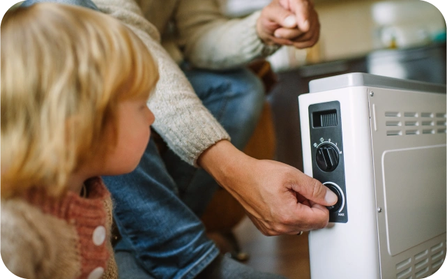 Persona, ģērbusies džemperī un džinsos, groza termostata slēdzi uz pārvietojama elektriskā sildītāja, priekšplāna mazs bērns džemperī vēro notiekošo.