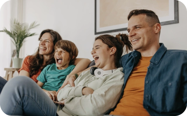 Četri cilvēki sēž cieši kopā uz dīvāna dzīvojamā istabā, fonā augs uz neliela galdiņa un ierāmēts attēls pie sienas.