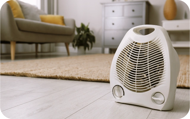Dzīvojamā istabā uz grīdas stāv balts siltā gaisa ventilators ar rokturi un diviem vadības slēdžiem; fonā bēšs paklājs, pelēks dīvāns ar krāsainiem spilveniem, pelēka kumode ar atvilktnēm un augs.