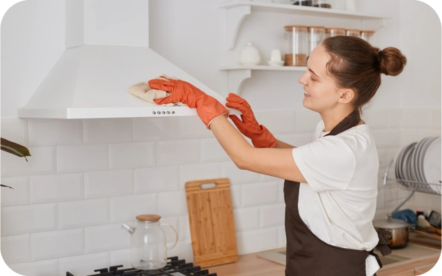 Sieviete ar oranžiem gumijas cimdiem un melnu priekšautu tīra baltu tvaika nosūcēju ar drānu; fonā balta flīžu siena un stikla tējkanna uz plīts.