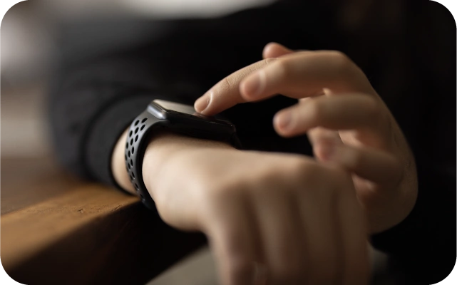 Tuvplāns ar personas roku, kas pieskaras viedpulkstenim ar melnu silikona siksniņu; fons izpludināts, izceļot darbību ar ierīci.