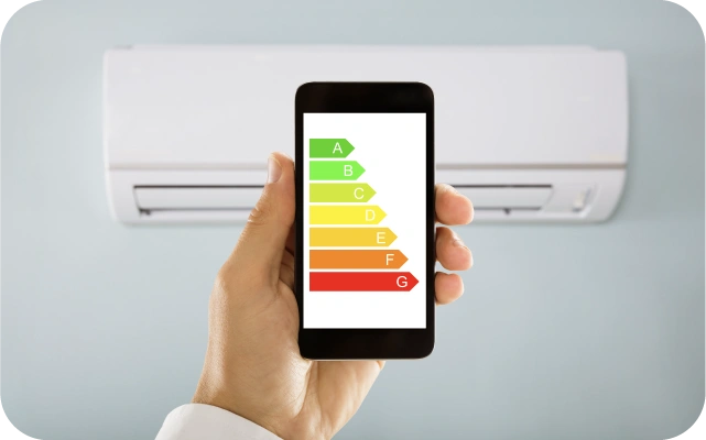 Cilvēka rokā ir mobilais telefons, kura ekrānā ir energoefektivitātes marķējums no A līdz G ar krāsainu skalu — no zaļa līdz sarkanam; fonā pie sienas piestiprināts gaisa kondicionieris.