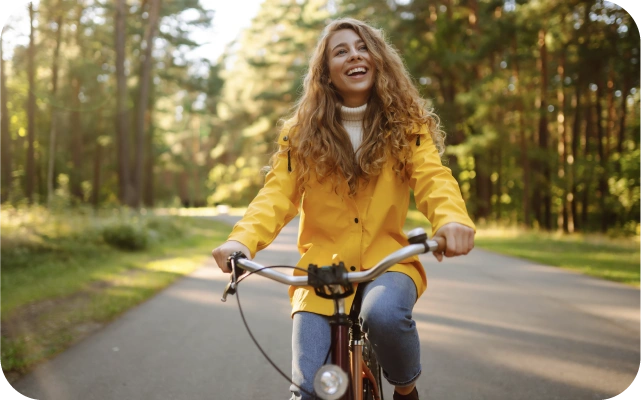 Sieviete dzeltenā vējjakā un zilās džinsās brauc ar velosipēdu pa asfaltētu taku, kas ved cauri zaļam mežam; saules stari filtrējas caur koku lapotni