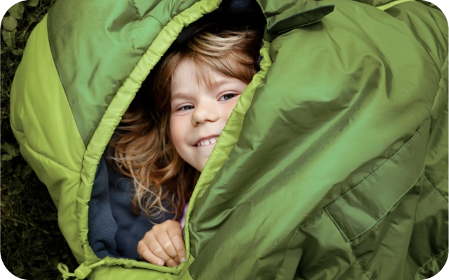 Bērns guļ zaļā guļammaisā zālē, atsegts viena roka, kas tur guļammaisa malu un galva.