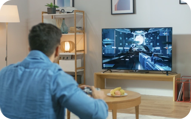 Vīrietis ar spēļu kontrolieri sēž uz dīvāna un spēlē videospēli uz liela televizora ekrāna, fonā koka kafijas galdiņš, grīdas lampa, plaukts ar dekoratīviem priekšmetiem, pie sienas ierāmēti attēli