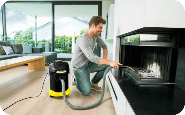 Vīrietis izmanto melni dzeltenu pelnu putekļu sūcēju, lai iztīrītu kamīnu — vīrietis tup un ar elastīgu cauruli savāc pelnus un gruži; fonā gaiša viesistaba ar dīvānu, koka kafijas galdiņu un lieliem logiem.