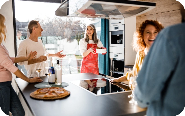 Moderna virtuve ar pieciem cilvēkiem, kas pulcējas ap virtuves salu — viens valkā sarkanu priekšautu un tur bļodu, uz virtuves salas virsmas ir pica, vīna pudele un glāze.