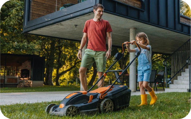 Bērns dzeltenos gumijas zābakos stumj zāles pļāvēja turoties pie roktura, kamēr pieaugušais sarkanā kreklā un zaļos šortos stāv blakus, palīdzot; viņi atrodas uz zāliena pie modernas mājas.