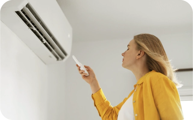 Sieviete dzeltenā kreklā ar tālvadības pulti rokā ieslēdz gaisa kondicionieri, kas atrodas viņai pretī pie sienas; fons gaišs.