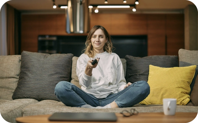 Sieviete baltā kreklā un džinsos sēž saliekusi kājas uz bēša dīvāna, rokā turot pulti. Uz dīvāna dzelteni, pelēki un tumši pelēki spilveni, priekšā koka kafijas galdiņš ar krūzīti, portatīvo datoru un brillēm.