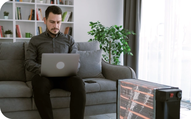 Vīrietis tumšās drēbēs sēž uz pelēka dīvāna ar portatīvo datoru klēpī, fonā grāmatu plaukts, augs podā un logs; priekšplānā novietots elektriskais sildītājs.