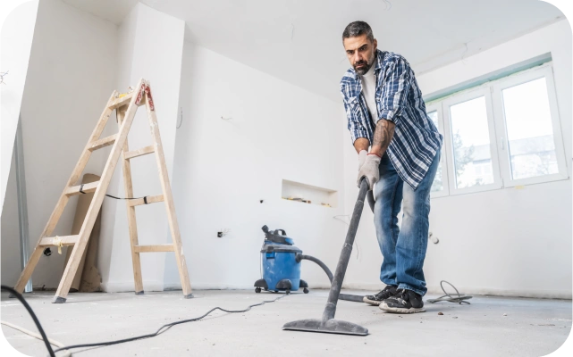 Vīrietis darba cimdos un zilā apģērbā tīra baltas elpas grīdu ar zilu celtniecības putekļu sūcēju; fonā logs, koka trepes un būvniecības piederumi.