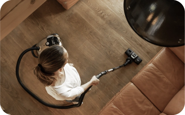 Sieviete gaišā apģērbā tīra koka grīdu pie brūna ādas dīvāna, izmantojot garu putekļu sūcēja šļūteni; skats no augšas.