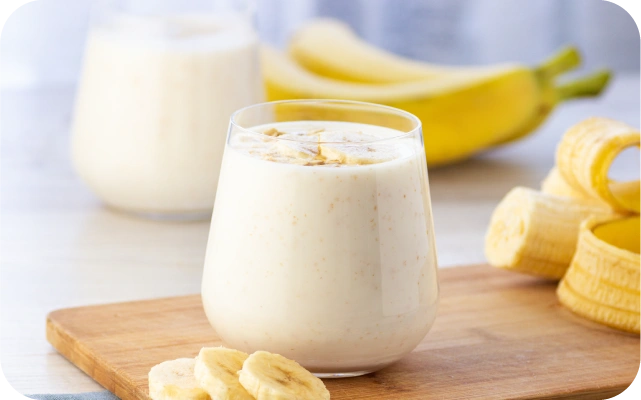 Glāze ar banānu smūtiju uz koka dēlīša, priekšplānā banāna šķēles, fonā veseli banāni un vēl viena glāze ar smūtiju.