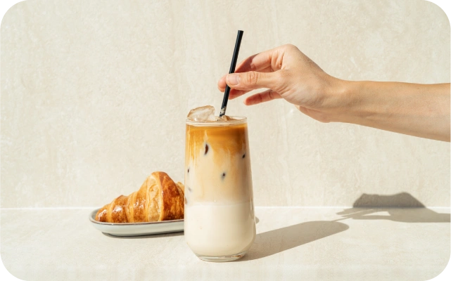 Cilvēka roka tur melnu salmiņu, kas atrodas glāzē ar ledus kafiju; blakus uz šķīvja ir kruasāns.