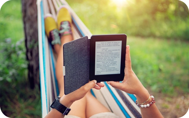 Cilvēks guļ krāsainā šūpuļtīklā ar e-grāmatu lasītāju rokās un lasa, fonā zaļa zāle un krūmi.