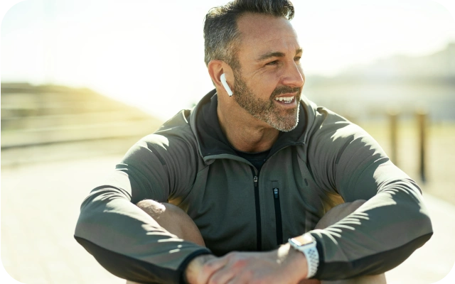 Vīrietis ar pelēku jaku un baltām bezvadu austiņām ausīs sēž ārā saulainā dienā.