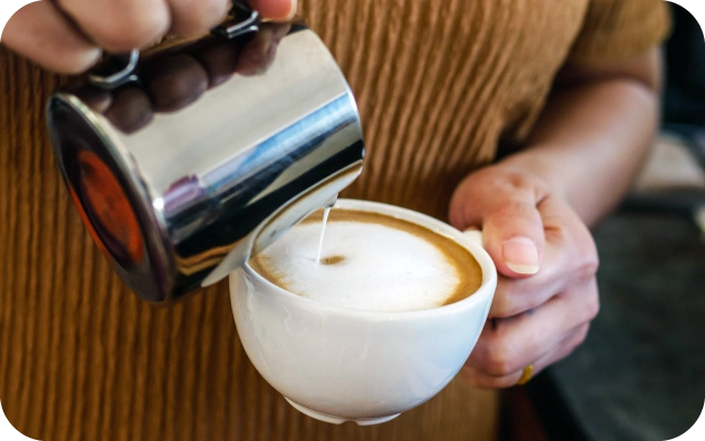 Tuvplāns ar piena ielešanu kafijā no metāla krūzes – latte ar piena putu kārtu.