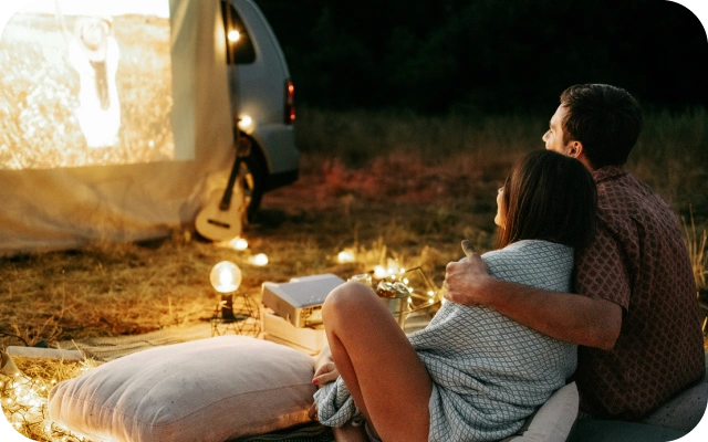 Pāris, sēžot dabā uz spilveniem, skatās filmu uz projektora ekrāna, kas atstutēts pret mašīnu, fonā zāliens, projektors un lampiņas.