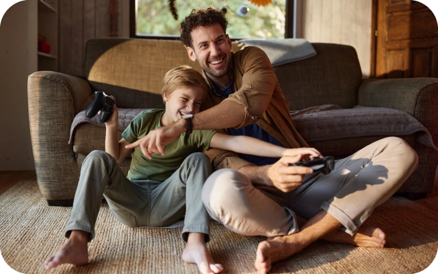 Divi smaidīgi cilvēki, pieaugušais un bērns, spēlē videospēles uz grīdas mājīgā dzīvojamā istabā atspieduši muguras pret pelēku dīvānu.