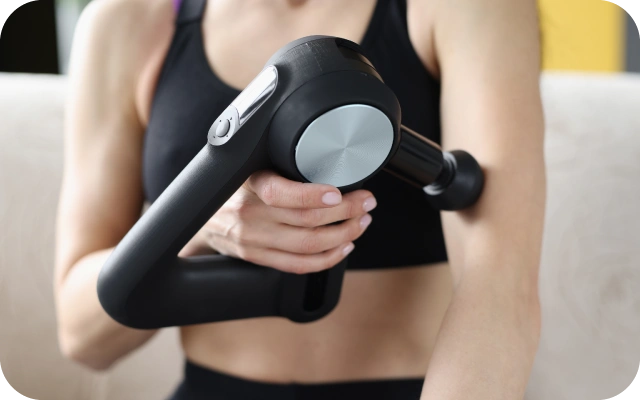 Sieviete melnā, sportiskā apģērbā lieto masāžas pistoli uz augšdelma – muskuļu atjaunošanai un sāpju mazināšanai.