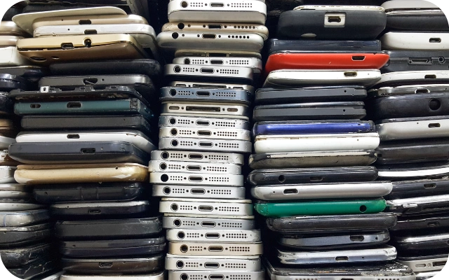 Liels daudzums vecu un nolietotu viedtālruņu sakrauti kaudzēs, redzami to apakšējie savienotāji, skaļruņu restes un nodiluši korpusi.