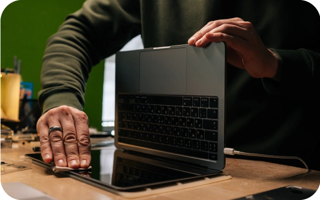 Cilvēks ar vienu roku tīra portatīvā datora ekrānu, bet ar otru roku pietur sagāzto datoru uz darbagalda. Fons rada darbnīcas atmosfēru.