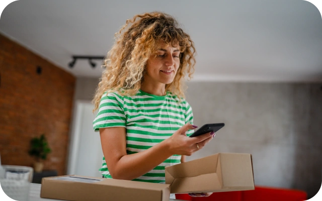 Sieviete tur rokā viedtālruni un piegādātu kartona kasti mājas vidē, kur redzami vairāki iepakojumi.