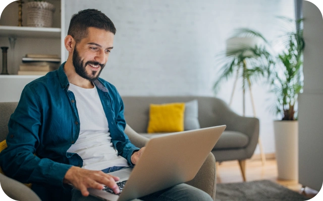 Vīrietis sēž viesistabā uz dīvāna ar portatīvo datoru klēpjī un skatās tajā. Fonā istabas augi un spilveni.