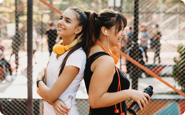 Divas meitenes sporta laukumā ar austiņām – viena meitene baltā T-kreklā ar dzeltenām austiņām apliktām ap kaklu, otra meitene melnā sporta kreklā ar baltām vadu austiņām ausīs; abām meitenēm rokās ūdens pudeles.