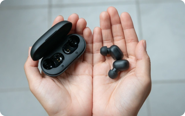 Melns bezvadu austiņu komplekts ar atvērtu uzlādes futrāli un austiņām cilvēka rokās.