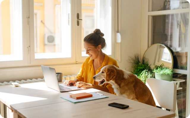 Sieviete dzeltenā kreklā strādā ar portatīvo datoru pie gaiša galda, blakus uz krēsla šēz suns, kas skatās ekrānā; uz galda atrodas piezīmju grāmatiņa, pildspalva un mobilais telefons.